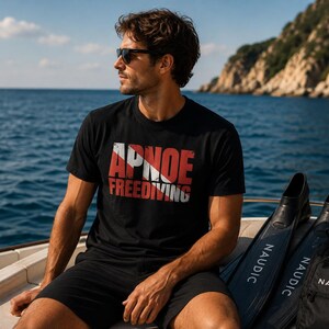 Puede incluir: Una camiseta negra de manga corta con el texto rojo y blanco APNOE FREEDIVING en un dise&ntilde;o gr&aacute;fico desgastado. La camiseta se combina con pantalones cortos negros y aletas de nataci&oacute;n negras de la marca Naudic junto a la prenda.