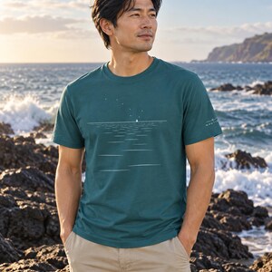 Peut inclure: Un t-shirt &agrave; manches courtes et col rond bleu sarcelle fonc&eacute; avec une impression graphique blanche repr&eacute;sentant une ligne d'horizon, un petit voilier et une constellation d'&eacute;toiles. La manche gauche porte un logo blanc avec le texte NAUTIVIBE.