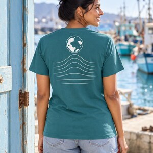 K&ouml;nnte beinhalten: Ein kurz&auml;rmeliges T-Shirt in Petrol mit einer wei&szlig;en Grafik auf der R&uuml;ckseite. Das Design besteht aus einer kreisf&ouml;rmigen Mondform &uuml;ber einer Reihe von f&uuml;nf horizontalen, wellenf&ouml;rmigen Linien, die nach unten hin breiter werden.