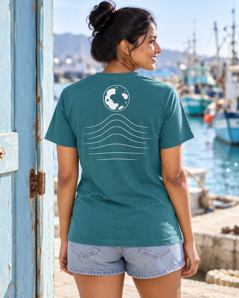 K&ouml;nnte beinhalten: Ein kurz&auml;rmeliges T-Shirt in Petrol mit einer wei&szlig;en Grafik auf der R&uuml;ckseite. Das Design besteht aus einer kreisf&ouml;rmigen Mondform &uuml;ber einer Reihe von f&uuml;nf horizontalen, wellenf&ouml;rmigen Linien, die nach unten hin breiter werden.
