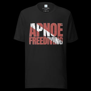 Puede incluir: Una camiseta negra de manga corta con el texto APNOE FREEDIVING en una fuente desgastada de color blanco y rojo. El dise&ntilde;o incorpora un patr&oacute;n de rayas blancas diagonales sobre las letras, imitando una bandera de buceo en el pecho.