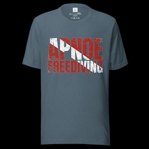 Puede incluir: Una camiseta de manga corta color azul pizarra con un estampado gr&aacute;fico desgastado en rojo y blanco. El dise&ntilde;o muestra el texto APNOE FREEDIVING superpuesto sobre un patr&oacute;n de rayas diagonales rojas y blancas, similar a una bandera de buceo.