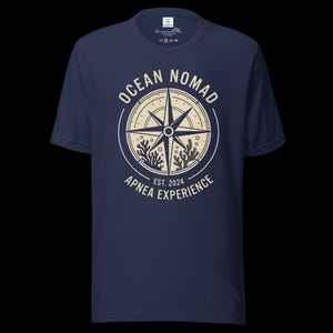 Op de afbeelding: Een marineblauw t-shirt met korte mouwen en een cr&egrave;mekleurige afbeelding van een kompasroos omringd door koraal, met de tekst "OCEAN NOMAD" en "APNEA EXPERIENCE EST. 2024" in een cirkelvormig ontwerp op het midden van de borst.