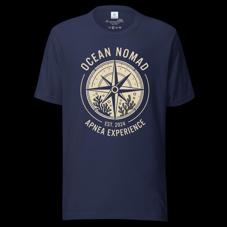 Op de afbeelding: Een marineblauw t-shirt met korte mouwen en een cr&egrave;mekleurige afbeelding van een kompasroos omringd door koraal, met de tekst "OCEAN NOMAD" en "APNEA EXPERIENCE EST. 2024" in een cirkelvormig ontwerp op het midden van de borst.