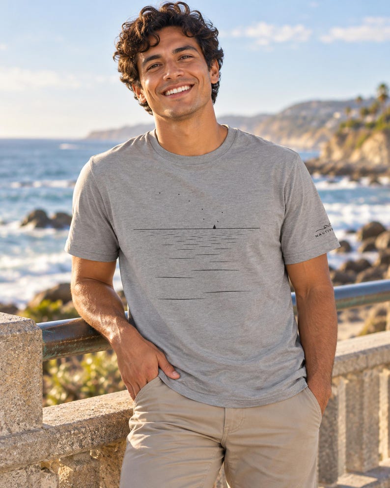 Peut inclure: Un t-shirt gris chin&eacute; &agrave; manches courtes orn&eacute; d'un graphisme minimaliste en lignes noires repr&eacute;sentant un voilier &agrave; l'horizon sous de petites &eacute;toiles. La manche porte un petit logo noir avec le texte NAUTIVE en police sans-serif.