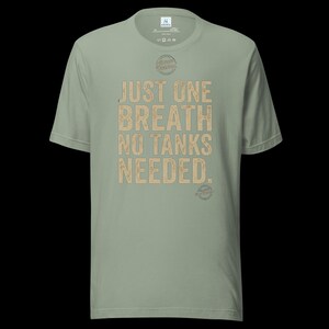 May include: A sage green short-sleeve t-shirt featuring the text "JUST ONE BREATH NO TANKS NEEDED." in a distressed tan font. The shirt includes a small circular logo above the text and a smaller circular logo near the bottom right hem.