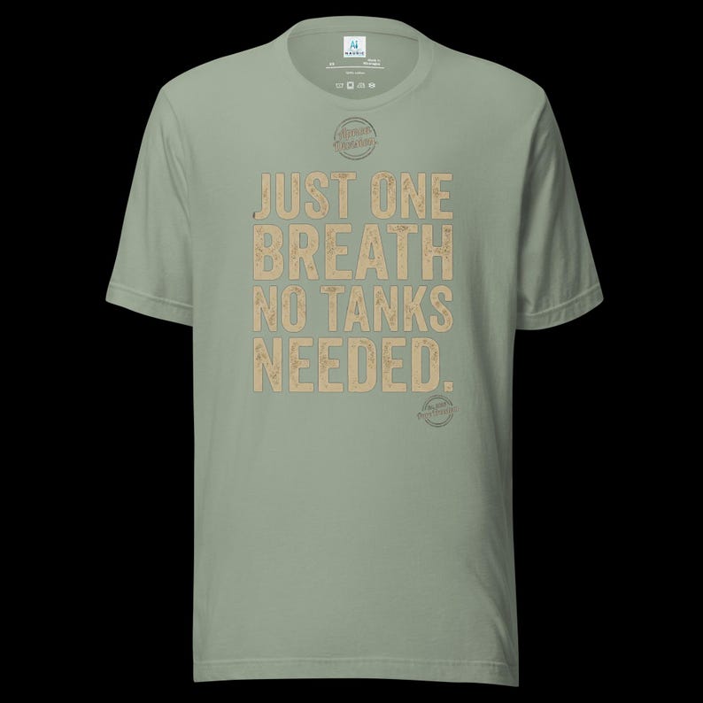 May include: A sage green short-sleeve t-shirt featuring the text "JUST ONE BREATH NO TANKS NEEDED." in a distressed tan font. The shirt includes a small circular logo above the text and a smaller circular logo near the bottom right hem.