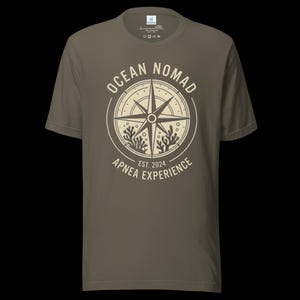 Op de afbeelding: Een olijfgroen t-shirt met korte mouwen en een ronde hals, voorzien van een cr&egrave;mekleurige opdruk op de borst. Het ontwerp toont een kompasroos omringd door koraalillustraties en de tekst "OCEAN NOMAD" en "APNEA EXPERIENCE EST. 2024".