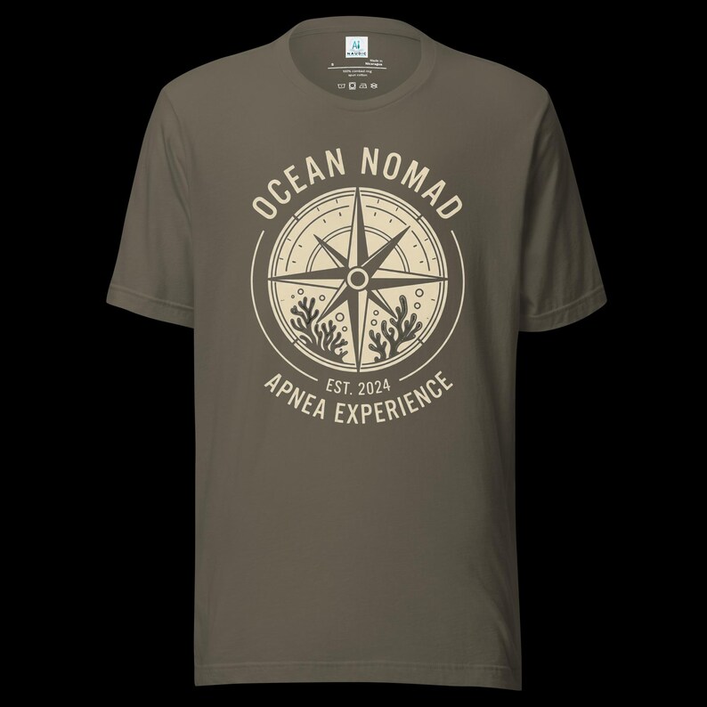 Op de afbeelding: Een olijfgroen t-shirt met korte mouwen en een ronde hals, voorzien van een cr&egrave;mekleurige opdruk op de borst. Het ontwerp toont een kompasroos omringd door koraalillustraties en de tekst "OCEAN NOMAD" en "APNEA EXPERIENCE EST. 2024".
