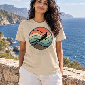 K&ouml;nnte beinhalten: Ein beiges T-Shirt mit einer kreisf&ouml;rmigen Grafik in einem Retro-Sonnenuntergangsdesign mit orangefarbenen, gelben und t&uuml;rkisfarbenen Streifen. Die Grafik zeigt die Silhouette eines Apnoetauchers mit Flossen und den Text "APNEA REBELLION EST. 2024 ONE KICK AT A TIME."
