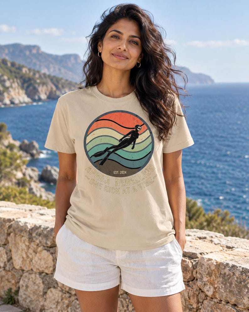 K&ouml;nnte beinhalten: Ein beiges T-Shirt mit einer kreisf&ouml;rmigen Grafik in einem Retro-Sonnenuntergangsdesign mit orangefarbenen, gelben und t&uuml;rkisfarbenen Streifen. Die Grafik zeigt die Silhouette eines Apnoetauchers mit Flossen und den Text "APNEA REBELLION EST. 2024 ONE KICK AT A TIME."
