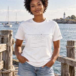 Peut inclure: Un t-shirt blanc &agrave; manches courtes avec un graphisme minimaliste en lignes noires repr&eacute;sentant un horizon, des ondulations d'eau et un petit voilier. Le texte NAUTIVE est imprim&eacute; en petites lettres noires sur la manche gauche.