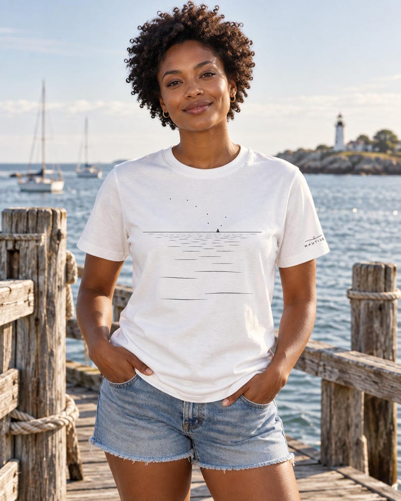 Peut inclure: Un t-shirt blanc &agrave; manches courtes avec un graphisme minimaliste en lignes noires repr&eacute;sentant un horizon, des ondulations d'eau et un petit voilier. Le texte NAUTIVE est imprim&eacute; en petites lettres noires sur la manche gauche.