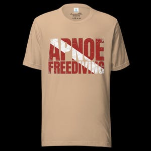 Puede incluir: Una camiseta de manga corta color canela con un estampado gr&aacute;fico desgastado en rojo y blanco en el pecho. El dise&ntilde;o muestra el texto APNOE y FREEDIVING superpuesto sobre una franja blanca diagonal, imitando el dise&ntilde;o de una bandera de buceo.