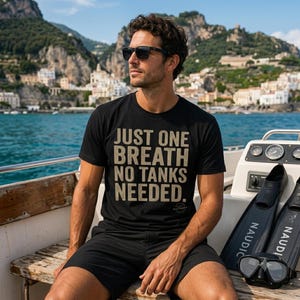 May include: A black short-sleeved t-shirt featuring the text "JUST ONE BREATH NO TANKS NEEDED." in a tan, distressed font. The shirt is worn with black shorts, black sunglasses, and is positioned near black diving fins and a diving mask.