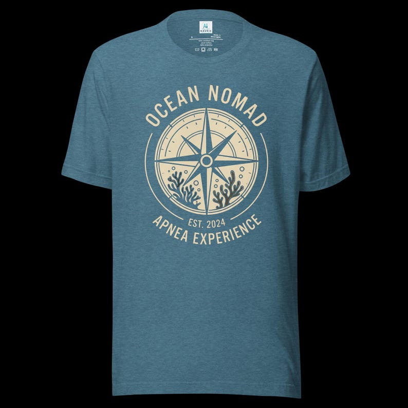 Op de afbeelding: Een gem&ecirc;leerd blauw t-shirt met korte mouwen en een cr&egrave;mekleurige cirkelvormige afbeelding op de borst. Het ontwerp bevat een kompasroos, koraalillustraties en de tekst "OCEAN NOMAD" en "APNEA EXPERIENCE EST. 2024" in een cirkelvormige lay-out.