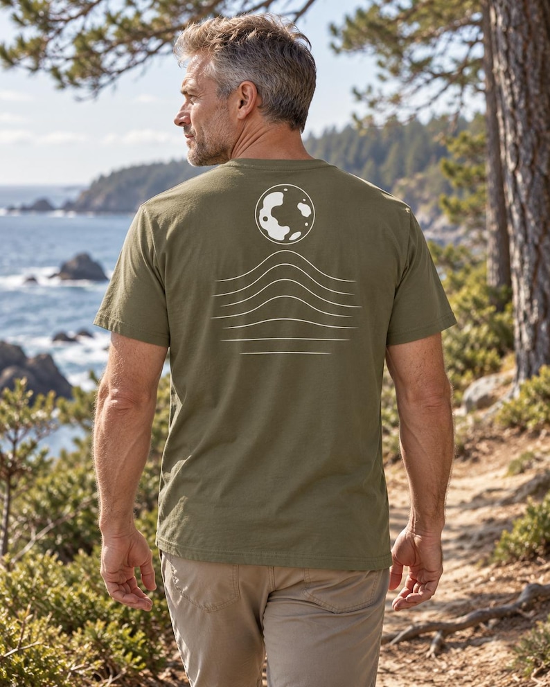 K&ouml;nnte beinhalten: Ein olivgr&uuml;nes Kurzarm-T-Shirt mit einer wei&szlig;en Grafik auf der R&uuml;ckseite. Das Design besteht aus einer kreisf&ouml;rmigen Mondform &uuml;ber einer Reihe von f&uuml;nf horizontalen, wellenf&ouml;rmigen Linien, die nach unten hin allm&auml;hlich flacher werden.