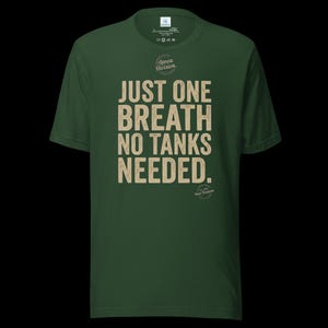 May include: A dark green short-sleeve t-shirt featuring the beige text "JUST ONE BREATH NO TANKS NEEDED." centered on the chest. Above the text is a circular logo with the words "Apnea Division." and below is a small circular emblem.