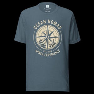Op de afbeelding: Een leiblauw t-shirt met korte mouwen en een cr&egrave;mekleurige cirkelvormige afbeelding op de borst. Het ontwerp bevat een kompasroos, koraalillustraties en de tekst "OCEAN NOMAD" en "APNEA EXPERIENCE EST. 2024" in een cirkelvormige lay-out.