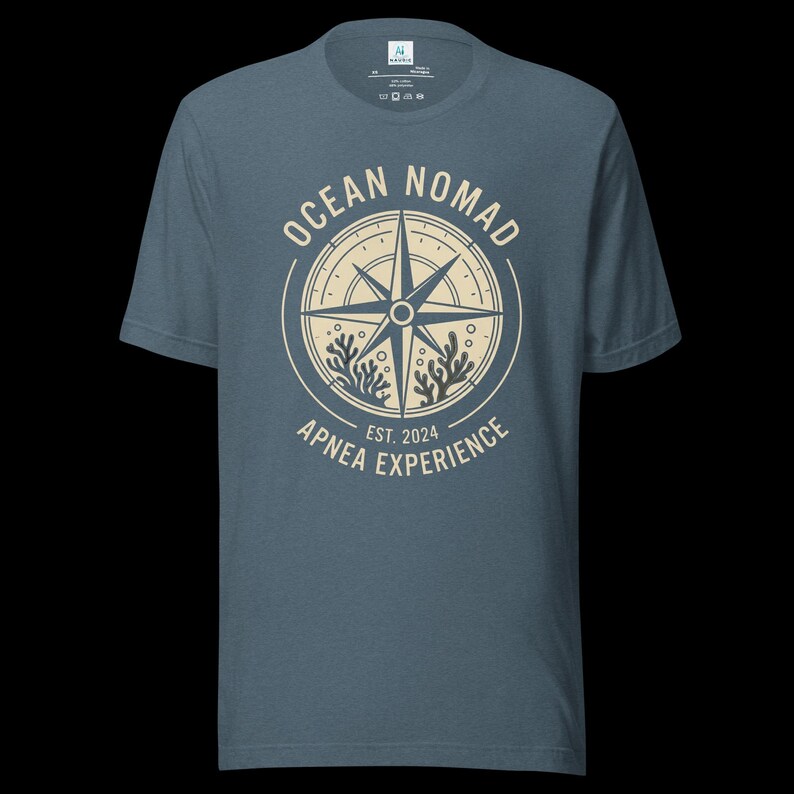 Op de afbeelding: Een leiblauw t-shirt met korte mouwen en een cr&egrave;mekleurige cirkelvormige afbeelding op de borst. Het ontwerp bevat een kompasroos, koraalillustraties en de tekst "OCEAN NOMAD" en "APNEA EXPERIENCE EST. 2024" in een cirkelvormige lay-out.