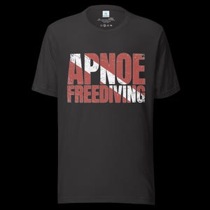 Puede incluir: Una camiseta gris carb&oacute;n de manga corta con el texto APNOE FREEDIVING en una fuente desgastada. Las letras est&aacute;n superpuestas con un patr&oacute;n de rayas diagonales rojas y blancas, que recuerda a una bandera de buceo, centrada en el pecho.
