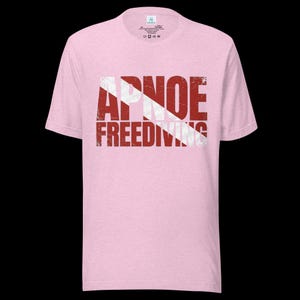 Puede incluir: Una camiseta de manga corta de color rosa claro con un estampado gr&aacute;fico desgastado en rojo y blanco en el pecho. El dise&ntilde;o incorpora un patr&oacute;n de rayas diagonales y el texto APNOE FREEDIVING en una fuente sans-serif negrita y desgastada.