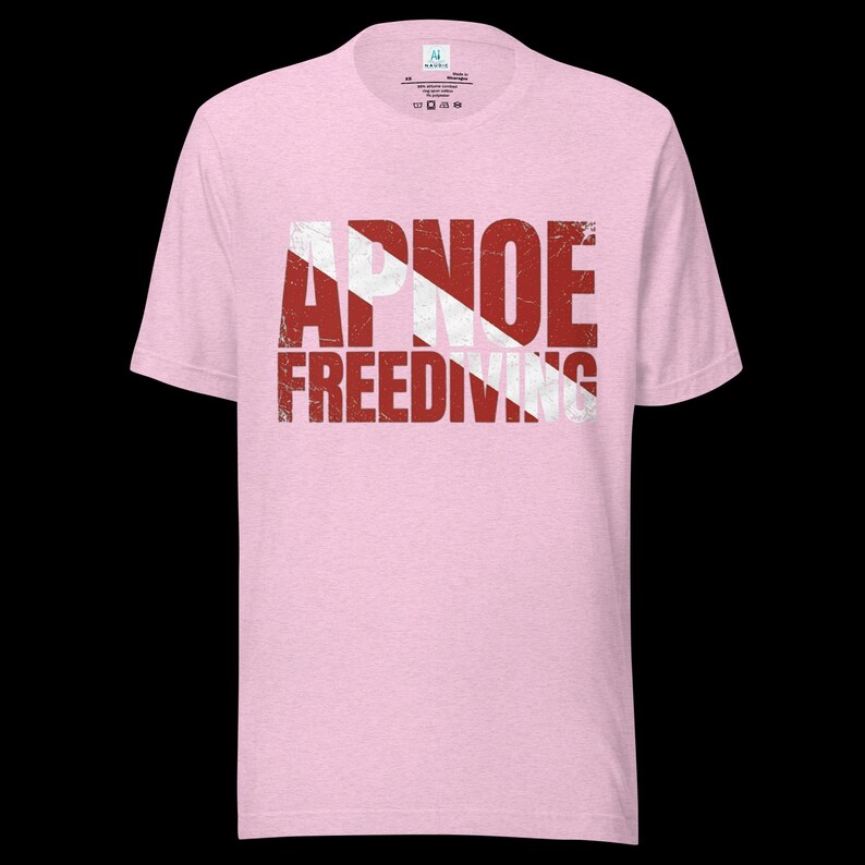 Puede incluir: Una camiseta de manga corta de color rosa claro con un estampado gr&aacute;fico desgastado en rojo y blanco en el pecho. El dise&ntilde;o incorpora un patr&oacute;n de rayas diagonales y el texto APNOE FREEDIVING en una fuente sans-serif negrita y desgastada.