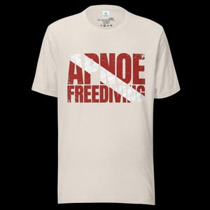 Puede incluir: Una camiseta de manga corta color crema con un gr&aacute;fico de bandera de buceo roja y blanca y el texto desgastado APNOE FREEDIVING superpuesto en el centro. La camiseta tiene cuello redondo y un corte est&aacute;ndar con una etiqueta interior visible.