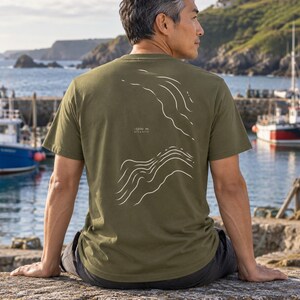 K&ouml;nnte beinhalten: Ein olivgr&uuml;nes Kurzarm-T-Shirt mit einer wei&szlig;en Liniengrafik auf der R&uuml;ckseite, die topografische Konturen darstellt. Der Text "-1200 m ATLANTIC" ist in kleinen wei&szlig;en Buchstaben zwischen den beiden Abschnitten des Wellenlinien-Designs gedruckt.