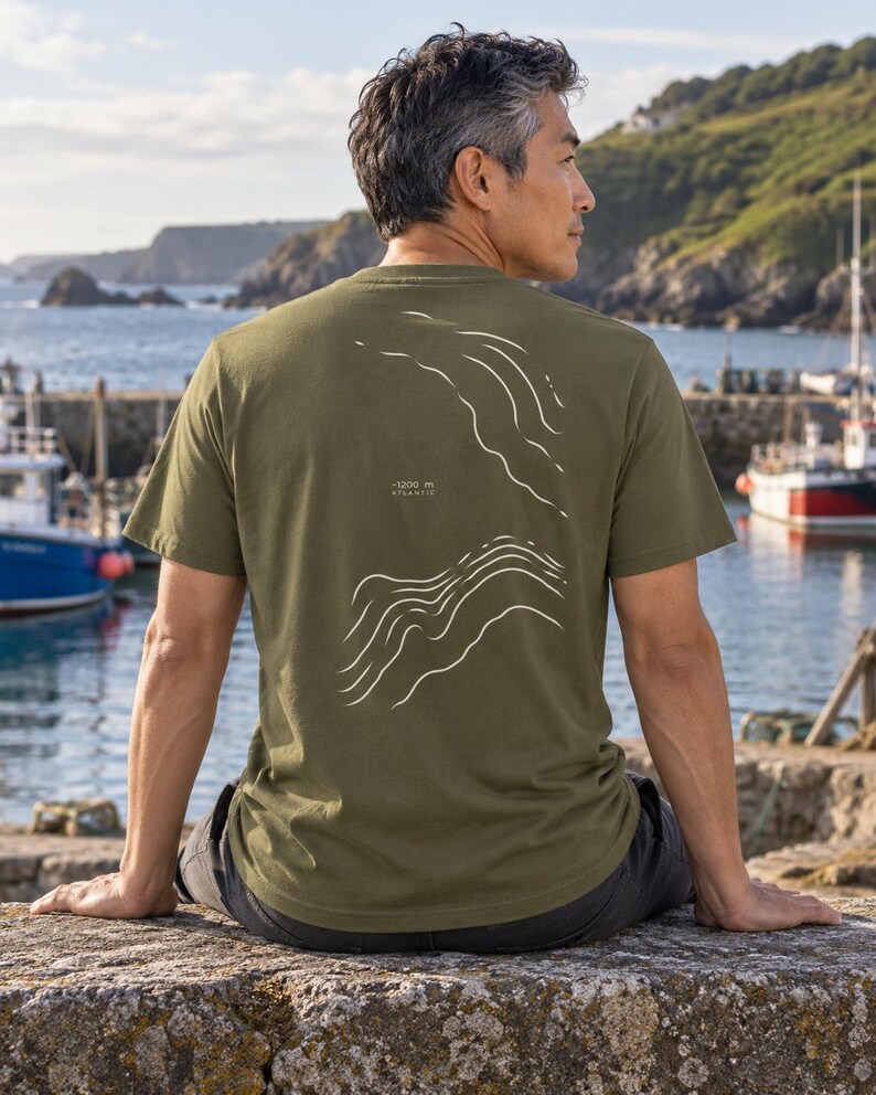 K&ouml;nnte beinhalten: Ein olivgr&uuml;nes Kurzarm-T-Shirt mit einer wei&szlig;en Liniengrafik auf der R&uuml;ckseite, die topografische Konturen darstellt. Der Text "-1200 m ATLANTIC" ist in kleinen wei&szlig;en Buchstaben zwischen den beiden Abschnitten des Wellenlinien-Designs gedruckt.