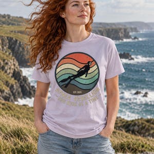 K&ouml;nnte beinhalten: Ein hellviolettes T-Shirt mit einem kreisf&ouml;rmigen Aufdruck, der ein Retro-Sonnenuntergangsdesign in orangefarbenen, gelben und t&uuml;rkisfarbenen Streifen zeigt. Eine Silhouette eines Apnoetauchers ist zentriert. Der Text lautet Apnea Rebellion, Est. 2024, One Kick At A Time.