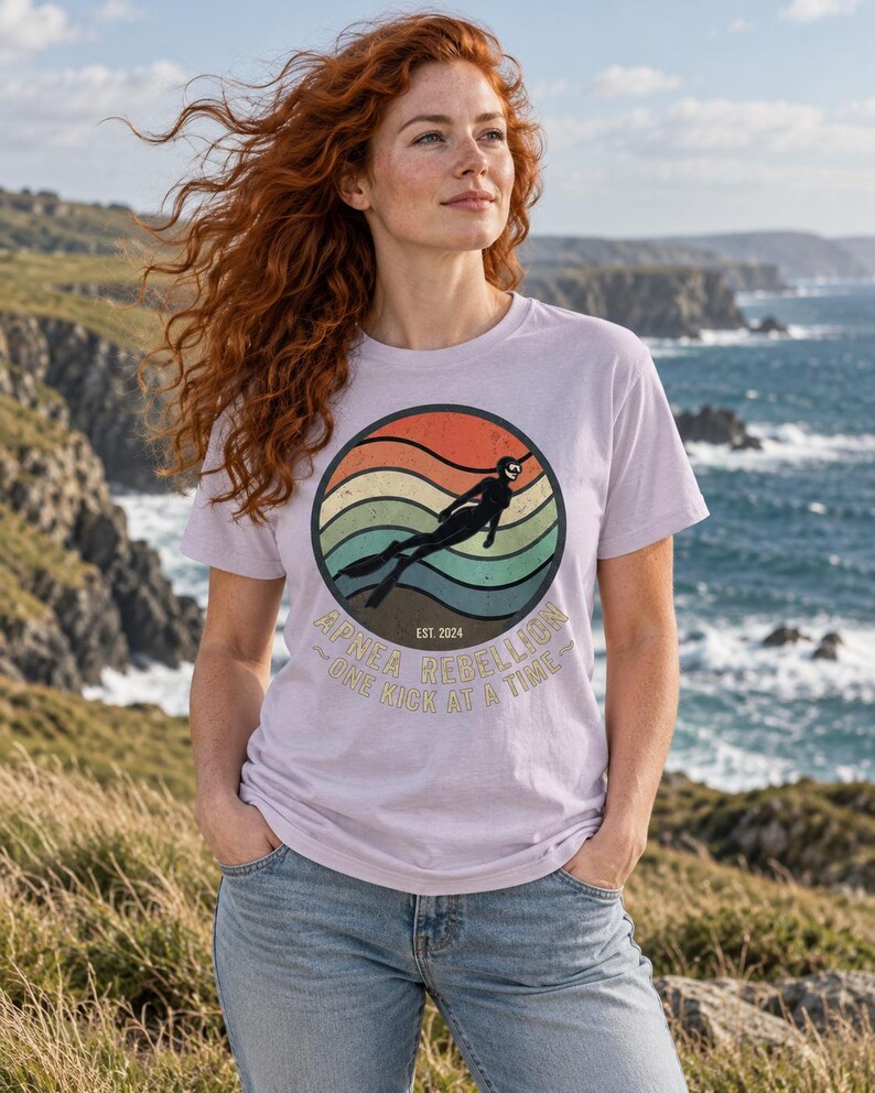 K&ouml;nnte beinhalten: Ein hellviolettes T-Shirt mit einem kreisf&ouml;rmigen Aufdruck, der ein Retro-Sonnenuntergangsdesign in orangefarbenen, gelben und t&uuml;rkisfarbenen Streifen zeigt. Eine Silhouette eines Apnoetauchers ist zentriert. Der Text lautet Apnea Rebellion, Est. 2024, One Kick At A Time.