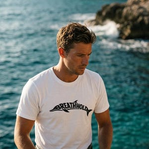 Może przedstawiać: Biały t-shirt z kr&oacute;tkim rękawem z czarną grafiką przedstawiającą sylwetkę delfina, w kt&oacute;rej korpus wkomponowano napis "BREATHHOLD". Koszulka posiada okrągły dekolt i wykonana jest z gładkiego materiału w jednolitym kolorze.