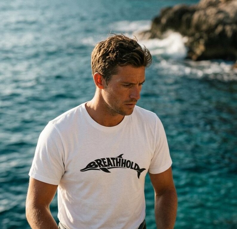 Może przedstawiać: Biały t-shirt z kr&oacute;tkim rękawem z czarną grafiką przedstawiającą sylwetkę delfina, w kt&oacute;rej korpus wkomponowano napis "BREATHHOLD". Koszulka posiada okrągły dekolt i wykonana jest z gładkiego materiału w jednolitym kolorze.