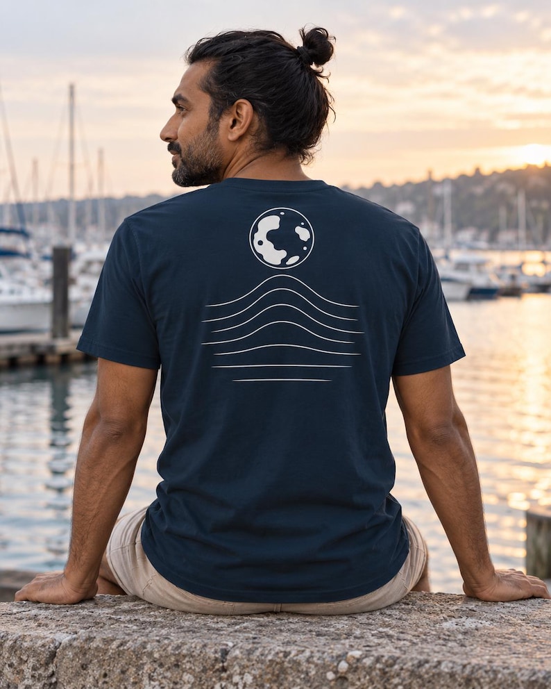 K&ouml;nnte beinhalten: Ein dunkelblaues Kurzarm-T-Shirt mit einer wei&szlig;en Grafik auf der R&uuml;ckseite. Das Design besteht aus einer kreisf&ouml;rmigen Mondform &uuml;ber einer Reihe von f&uuml;nf horizontalen, wellenf&ouml;rmigen Linien, die zum unteren Rand des Drucks hin flacher werden.