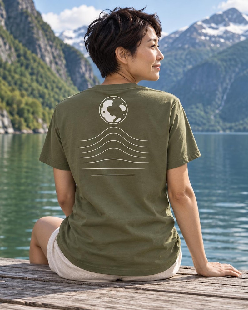 K&ouml;nnte beinhalten: Ein olivgr&uuml;nes Kurzarm-T-Shirt mit einer wei&szlig;en Grafik auf der R&uuml;ckseite. Das Design besteht aus einer kreisf&ouml;rmigen Mondform &uuml;ber einer Reihe von f&uuml;nf horizontalen Wellenlinien, die zum unteren Rand des Drucks hin flacher werden.