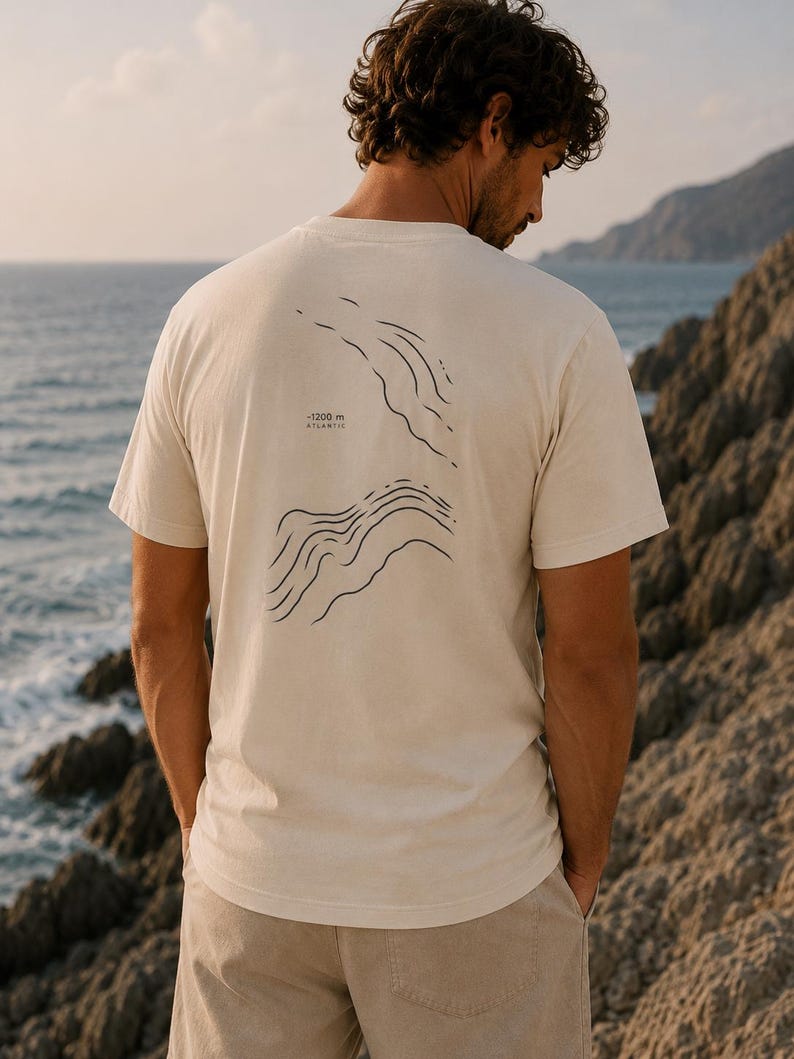 Könnte beinhalten: Ein cremefarbenes Kurzarm-T-Shirt mit einer schwarzen Linien-Grafik auf der Rückseite. Das Design zeigt abstrakte topografische Wellenmuster und enthält den aufgedruckten Text -1200 m ATLANTIC. Das T-Shirt hat einen Rundhalsausschnitt.