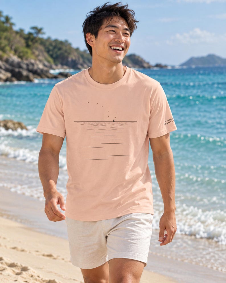 Peut inclure: Un t-shirt &agrave; manches courtes de couleur p&ecirc;che avec un graphisme au trait noir repr&eacute;sentant une ligne d'horizon, un petit voilier et une s&eacute;rie de points &eacute;voquant des oiseaux. La manche porte un petit logo noir avec le texte NAUTICA.
