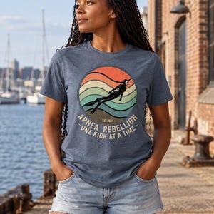 K&ouml;nnte beinhalten: Ein blau meliertes Kurzarm-T-Shirt mit einer kreisf&ouml;rmigen Grafik, die ein Retro-Sonnenuntergangsdesign, die Silhouette eines Apnoetauchers und den Text "APNEA REBELLION EST. 2024 ONE KICK AT A TIME" in hellem Beige auf der Vorderseite zeigt.