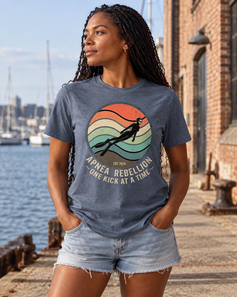 K&ouml;nnte beinhalten: Ein blau meliertes Kurzarm-T-Shirt mit einer kreisf&ouml;rmigen Grafik, die ein Retro-Sonnenuntergangsdesign, die Silhouette eines Apnoetauchers und den Text "APNEA REBELLION EST. 2024 ONE KICK AT A TIME" in hellem Beige auf der Vorderseite zeigt.