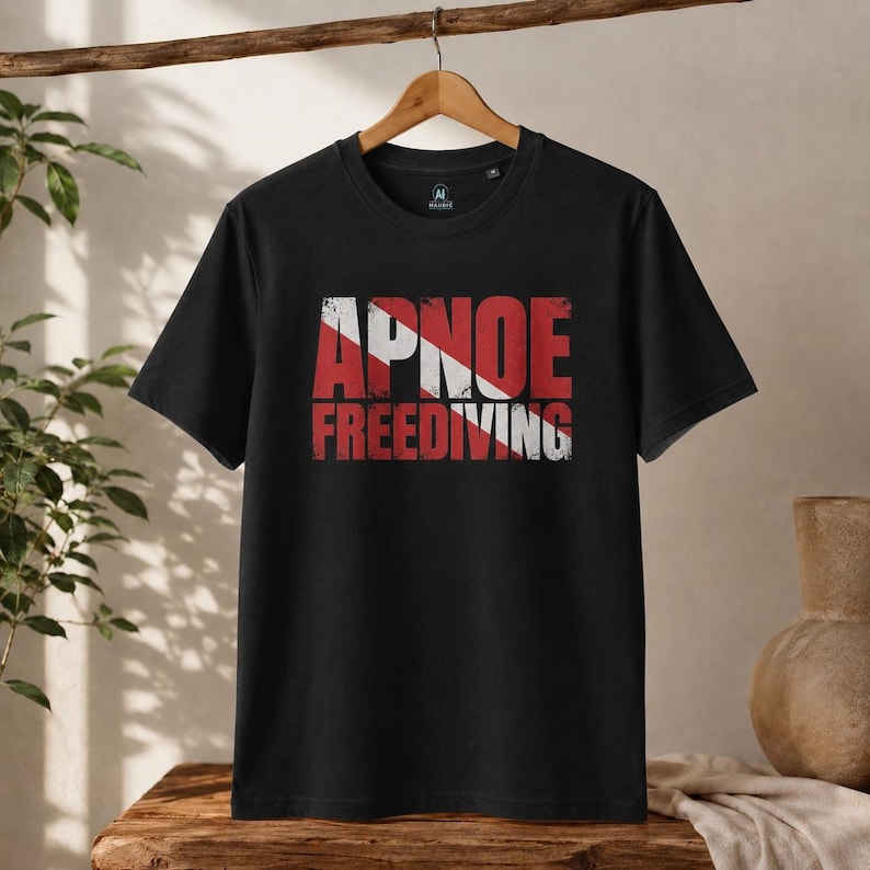 Könnte beinhalten: Ein schwarzes kurzärmeliges T-Shirt mit einem Grafikdruck auf der Brust. Das Design zeigt das Wort APNOE über dem Wort FREEDIVING in einer abgenutzten roten Schrift, überlagert von einem weißen diagonalen Streifen, der an eine Taucherflagge erinnert.