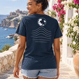 K&ouml;nnte beinhalten: Ein dunkelblaues T-Shirt mit kurzen &Auml;rmeln und einer wei&szlig;en Grafik auf der R&uuml;ckseite. Das Design besteht aus einer kreisf&ouml;rmigen Mondform &uuml;ber einer Reihe von f&uuml;nf horizontalen, wellenf&ouml;rmigen Linien, die nach unten hin k&uuml;rzer werden.