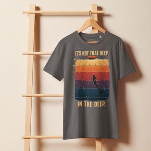 May include: A charcoal gray t-shirt featuring a graphic print of a diver underwater with a heart rate monitor line overlay. The text reads IT'S NOT THAT DEEP. IN THE DEEP. The design includes depth markers from 0m to 50m in a vertical color gradient.