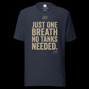 May include: A dark navy blue short-sleeve t-shirt featuring the beige text JUST ONE BREATH NO TANKS NEEDED. centered on the chest. Above the text is a circular logo with the words Apnea Division, and below is a small circular logo with Pure Freedom.