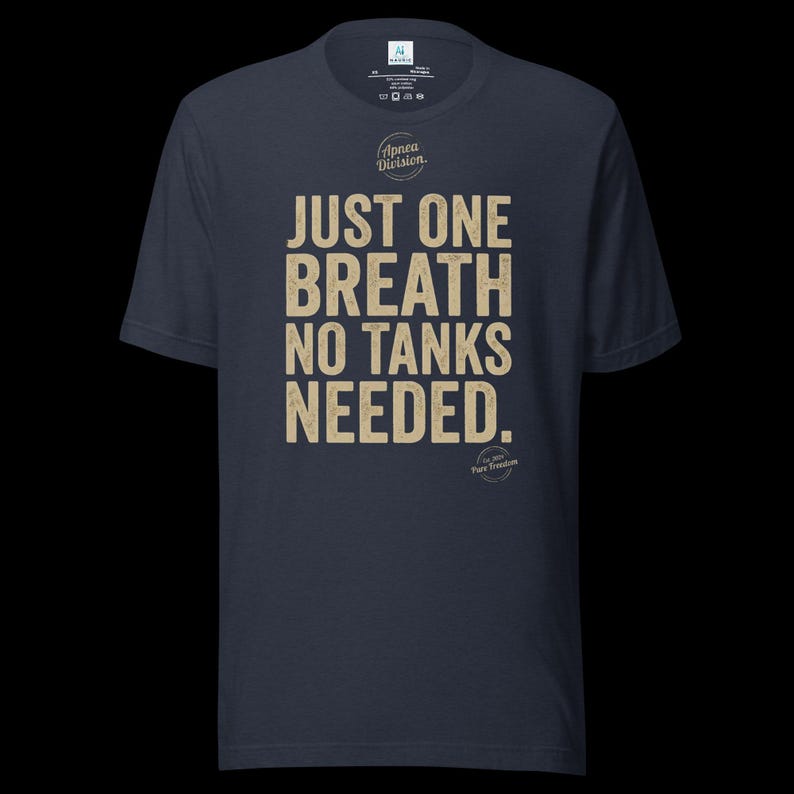 May include: A dark navy blue short-sleeve t-shirt featuring the beige text JUST ONE BREATH NO TANKS NEEDED. centered on the chest. Above the text is a circular logo with the words Apnea Division, and below is a small circular logo with Pure Freedom.