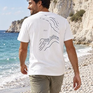 K&ouml;nnte beinhalten: Ein wei&szlig;es Kurzarm-T-Shirt mit einem dunkelblauen Grafikdruck topografischer H&ouml;henlinien auf der R&uuml;ckseite. Das Design enth&auml;lt den Text -1200 m ATLANTIC, der in kleinen schwarzen Buchstaben zwischen den beiden wellenf&ouml;rmigen Linienmustern gedruckt ist.