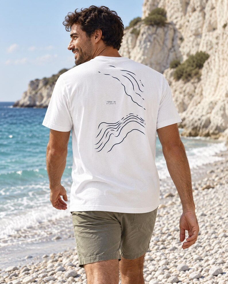 K&ouml;nnte beinhalten: Ein wei&szlig;es Kurzarm-T-Shirt mit einem dunkelblauen Grafikdruck topografischer H&ouml;henlinien auf der R&uuml;ckseite. Das Design enth&auml;lt den Text -1200 m ATLANTIC, der in kleinen schwarzen Buchstaben zwischen den beiden wellenf&ouml;rmigen Linienmustern gedruckt ist.
