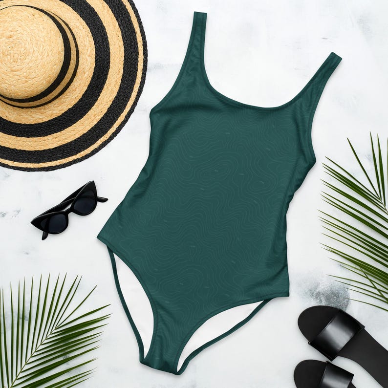 Peut inclure: Un maillot de bain une pi&egrave;ce vert fonc&eacute; avec un motif topographique subtil, accompagn&eacute; d'un chapeau de paille &agrave; ruban noir, de lunettes de soleil &oelig;il-de-chat noires et de sandales noires, le tout dispos&eacute; avec des feuilles de palmier.
