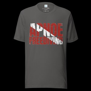 Puede incluir: Una camiseta de manga corta gris oscuro con un estampado gr&aacute;fico desgastado en el pecho. El dise&ntilde;o muestra el texto APNOE FREEDIVING en una fuente negrita y desgastada, superpuesto con un patr&oacute;n de rayas diagonales rojas y blancas.