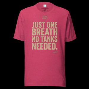 May include: A heathered red short-sleeve t-shirt featuring the text "JUST ONE BREATH NO TANKS NEEDED." printed in a distressed beige font on the chest. The shirt includes a small circular logo above the text and a brand label at the inner neck.